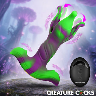 A green and purple silicone adult novelty device shown floating above a rock in a fantasy forest setting, with a small black remote control labeled CREATURE COCKS in the lower right. The image includes the text 'CREATURE COCKS' at the bottom.