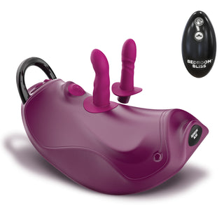 A purple and magenta dual-stimulation personal massager with a black carrying handle, shown alongside its black wireless remote control labeled 'BEDROOM BLISS'.