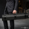 A man in a dark gray suit holds a long, black Master Series bondage equipment case with a textured fabric exterior and multiple black straps and buckles.