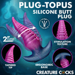 Promotional graphic for the Plug-Topus Silicone Butt Plug by Creature Cocks, showing a 2-inch tapered butt plug with a pink-to-purple gradient and suction-stripe texture. The graphic includes circular insets highlighting the tapered tip, stimulation texture, and ergonomic base, set against a dark teal background with text detailing product features.