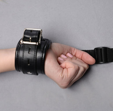Close-up of padded black wrist restraint cuff with silver buckle hardware connected to adjustable bed strap