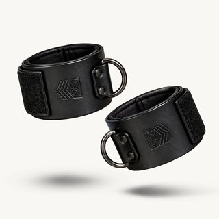 Pair of black Leather Wrist Cuffs closed with Velcro and metal D-rings on a white background