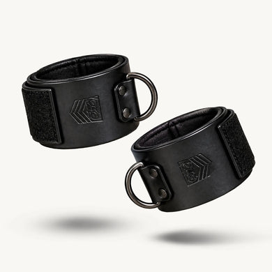 Pair of black Leather Wrist Cuffs closed with Velcro and metal D-rings on a white background