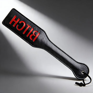 Black imprint spanking paddle with red BITCH lettering, glossy leather-look surface with wrist loop for impact play