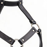 Close-up detail of black leather harness bra straps with silver O-rings, rivets, and smooth faux leather texture