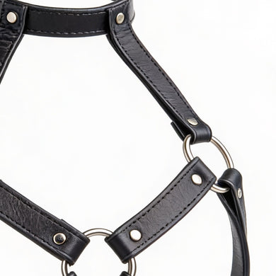 Close-up detail of black leather harness bra straps with silver O-rings, rivets, and smooth faux leather texture